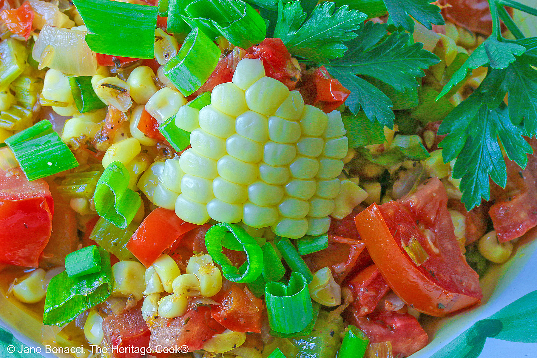 Close up of the corn in the bowl; A bowl with a blue and green decorated rim filled with the yellow and red Fresh Corn and Tomato Salad with a piece of corn from the cob on top and a whole tomato in front sitting on a vivid blue cloth © 2026 Jane Bonacci, The Heritage Cook.