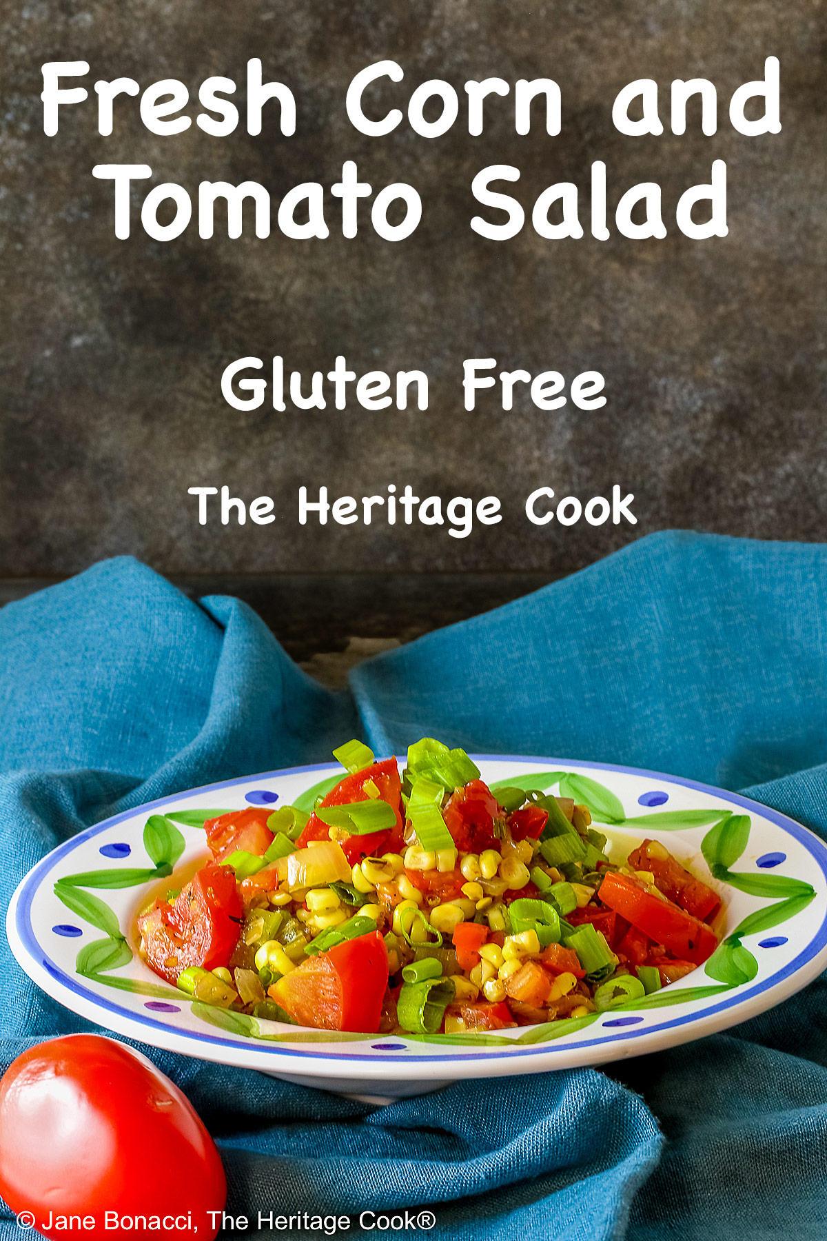 A bowl with a blue and green decorated rim filled with the yellow and red Fresh Corn and Tomato Salad with a piece of corn from the cob on top and a whole tomato in front sitting on a vivid blue cloth © 2026 Jane Bonacci, The Heritage Cook.