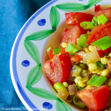 A bowl with a blue and green decorated rim filled with the yellow and red Fresh Corn and Tomato Salad with a piece of corn from the cob on top and a whole tomato in front sitting on a vivid blue cloth © 2026 Jane Bonacci, The Heritage Cook.