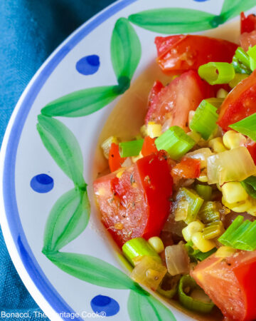A bowl with a blue and green decorated rim filled with the yellow and red Fresh Corn and Tomato Salad with a piece of corn from the cob on top and a whole tomato in front sitting on a vivid blue cloth © 2026 Jane Bonacci, The Heritage Cook.