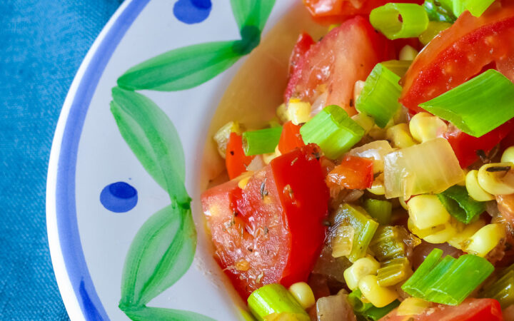 A bowl with a blue and green decorated rim filled with the yellow and red Fresh Corn and Tomato Salad with a piece of corn from the cob on top and a whole tomato in front sitting on a vivid blue cloth © 2026 Jane Bonacci, The Heritage Cook.
