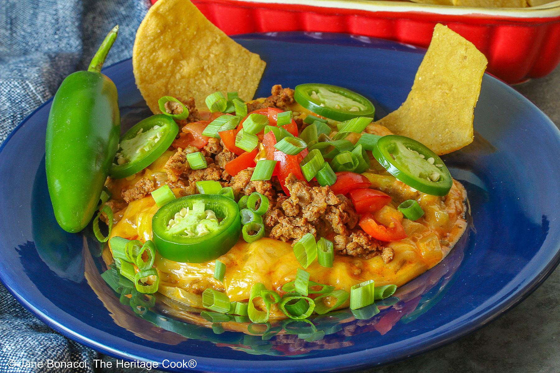 Dark blue bowl filled with the golden/orange Queso con Chorizo dip, topped with cooked chorizo, chopped tomatoes, jalapeno peppers, and sliced green onions with two tortilla chips tucked in beside the dip, sitting in front of a red rectangular dish overflowing with tortilla chips, with a blue cloth behind the dishes © 2026 Jane Bonacci, The Heritage Cook.