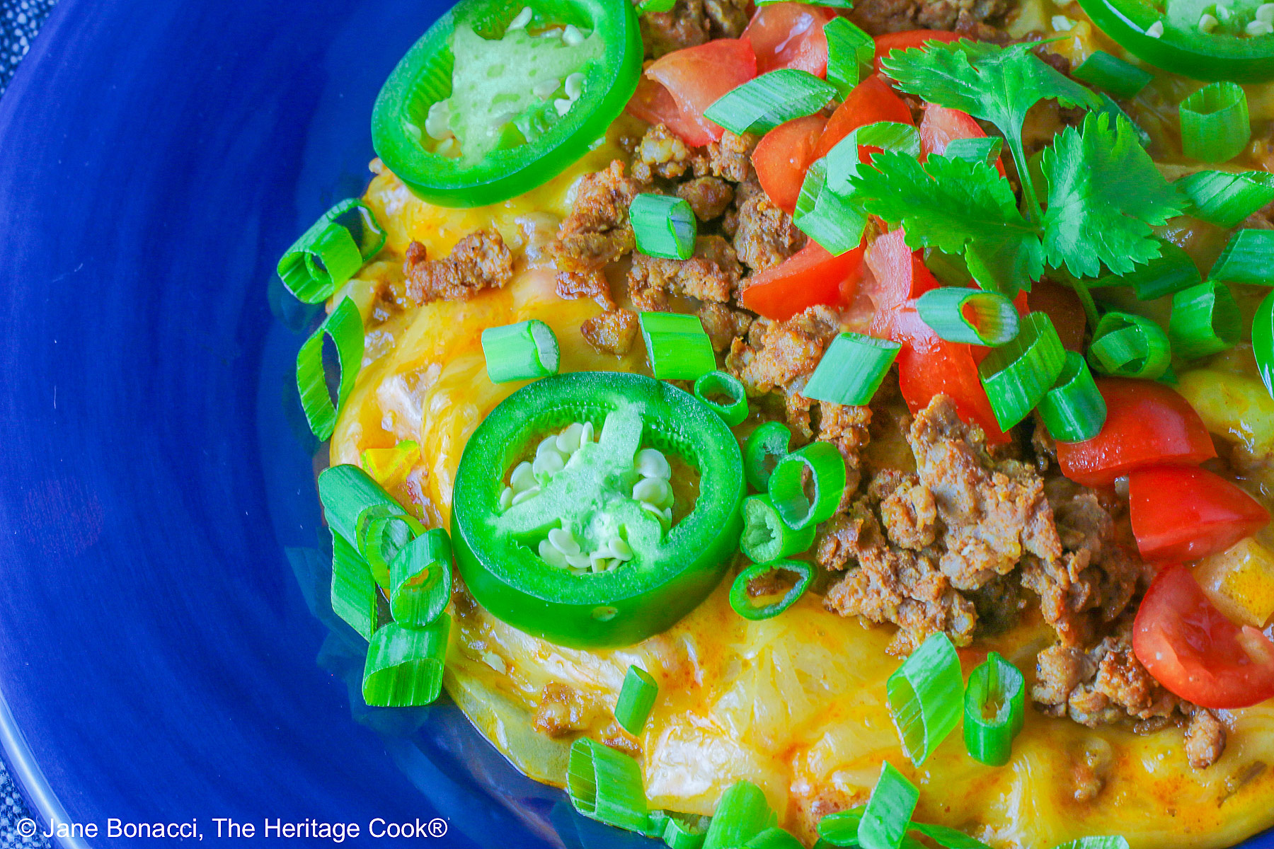 Dark blue bowl filled with the golden/orange Queso con Chorizo dip, topped with cooked chorizo, chopped tomatoes, jalapeno peppers, and sliced green onions with two tortilla chips tucked in beside the dip, sitting in front of a red rectangular dish overflowing with tortilla chips, with a blue cloth behind the dishes © 2026 Jane Bonacci, The Heritage Cook.