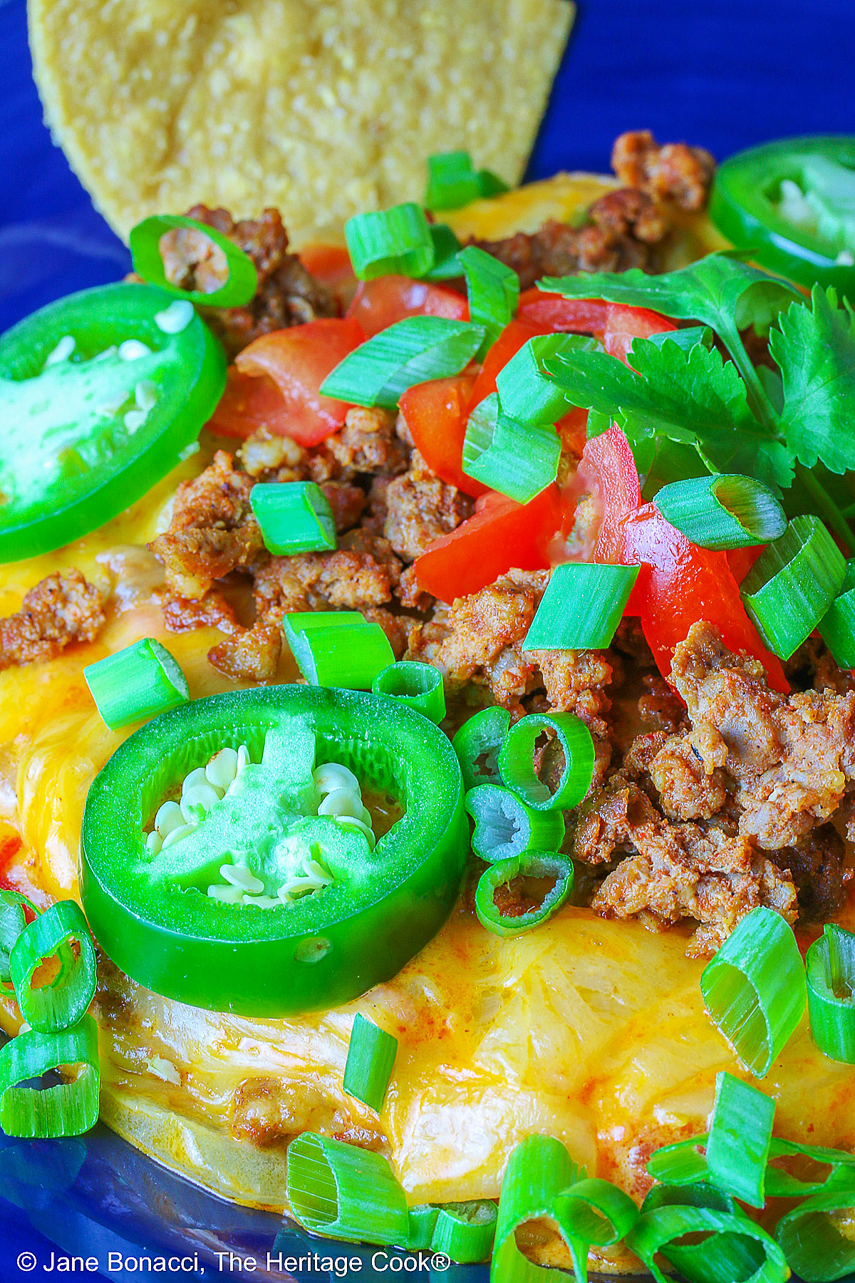 Dark blue bowl filled with the golden/orange Queso con Chorizo dip, topped with cooked chorizo, chopped tomatoes, jalapeno peppers, and sliced green onions with two tortilla chips tucked in beside the dip, sitting in front of a red rectangular dish overflowing with tortilla chips, with a blue cloth behind the dishes © 2026 Jane Bonacci, The Heritage Cook.