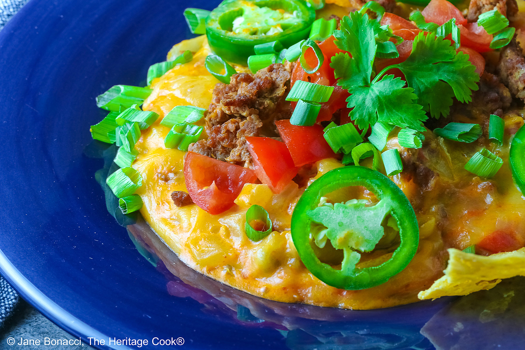 Dark blue bowl filled with the golden/orange Queso con Chorizo dip, topped with cooked chorizo, chopped tomatoes, jalapeno peppers, and sliced green onions with two tortilla chips tucked in beside the dip, sitting in front of a red rectangular dish overflowing with tortilla chips, with a blue cloth behind the dishes © 2026 Jane Bonacci, The Heritage Cook.