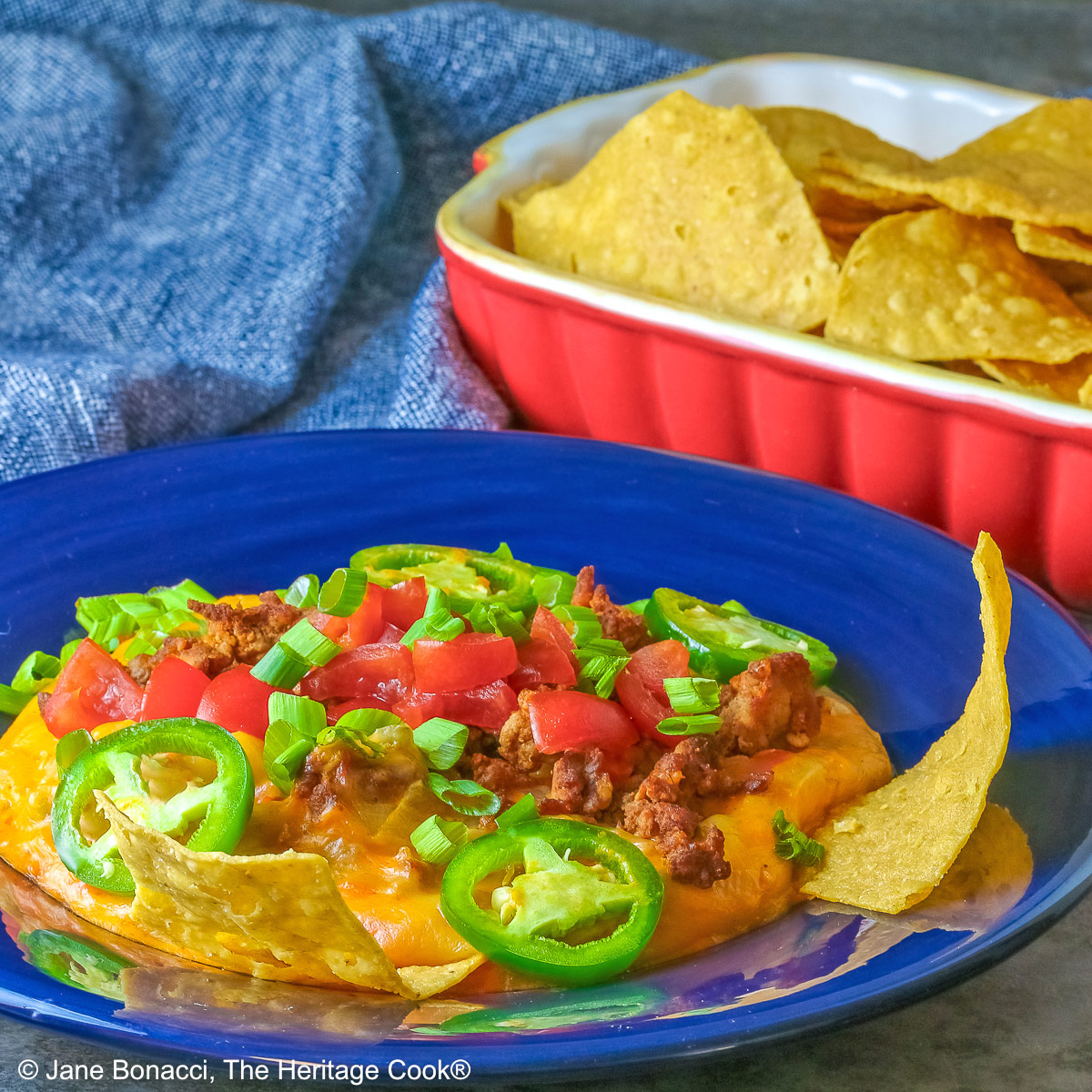 Dark blue bowl filled with the golden/orange Queso con Chorizo dip, topped with cooked chorizo, chopped tomatoes, jalapeno peppers, and sliced green onions with two tortilla chips tucked in beside the dip, sitting in front of a red rectangular dish overflowing with tortilla chips, with a blue cloth behind the dishes © 2026 Jane Bonacci, The Heritage Cook.