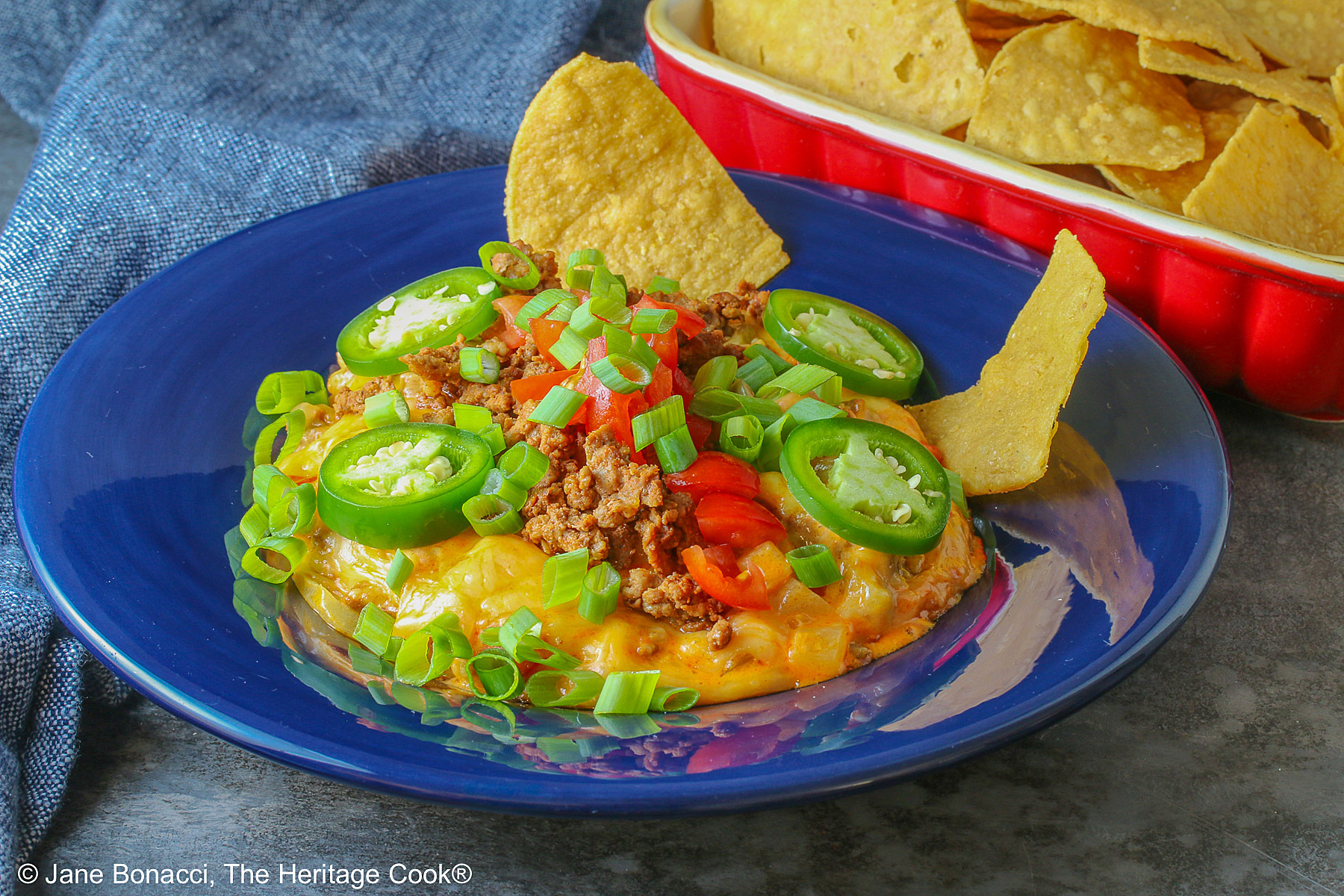 Dark blue bowl filled with the golden/orange Queso con Chorizo dip, topped with cooked chorizo, chopped tomatoes, jalapeno peppers, and sliced green onions with two tortilla chips tucked in beside the dip, sitting in front of a red rectangular dish overflowing with tortilla chips, with a blue cloth behind the dishes © 2026 Jane Bonacci, The Heritage Cook.
