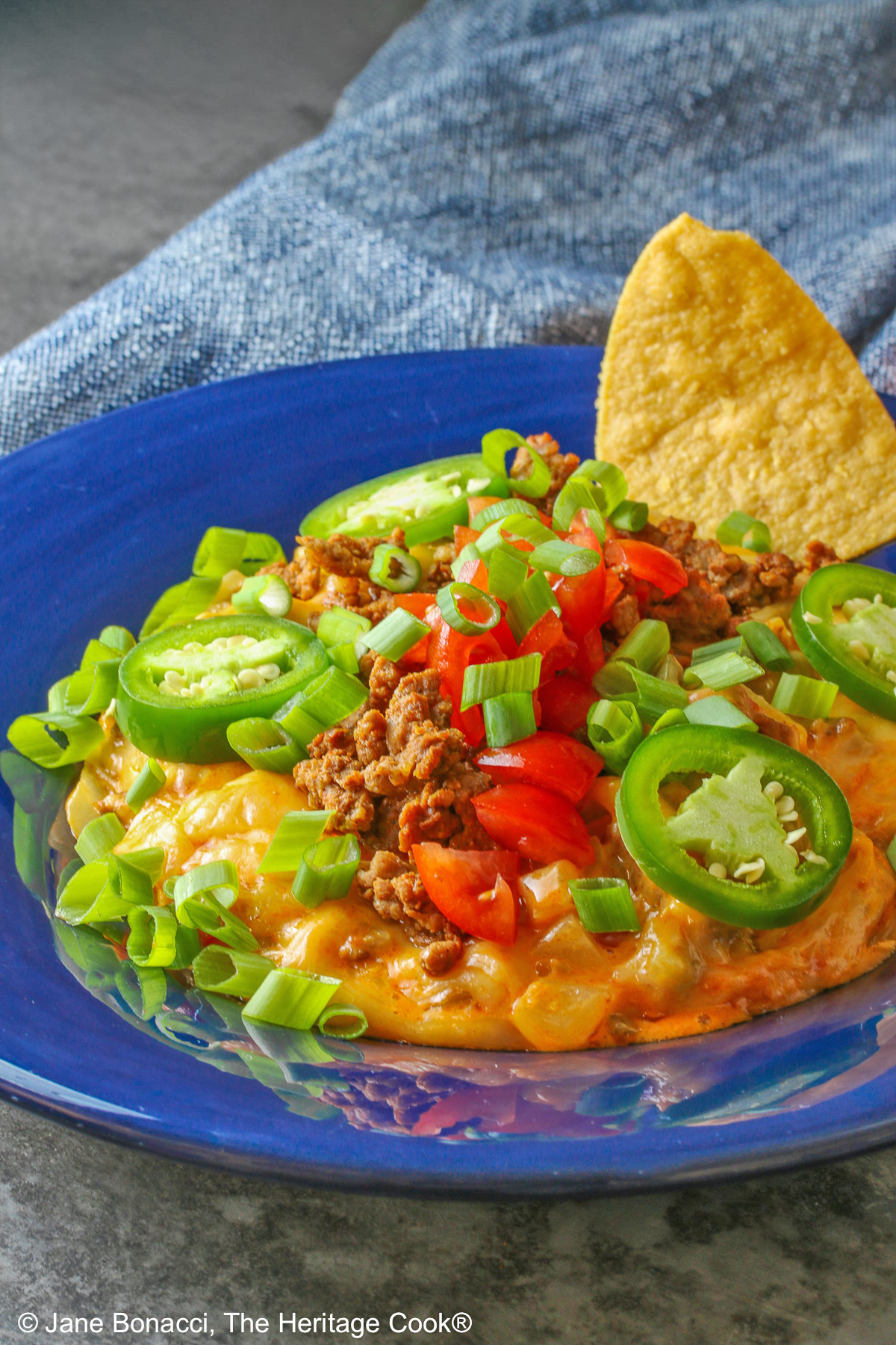 Dark blue bowl filled with the golden/orange Queso con Chorizo dip, topped with cooked chorizo, chopped tomatoes, jalapeno peppers, and sliced green onions with two tortilla chips tucked in beside the dip, sitting in front of a red rectangular dish overflowing with tortilla chips, with a blue cloth behind the dishes © 2026 Jane Bonacci, The Heritage Cook.
