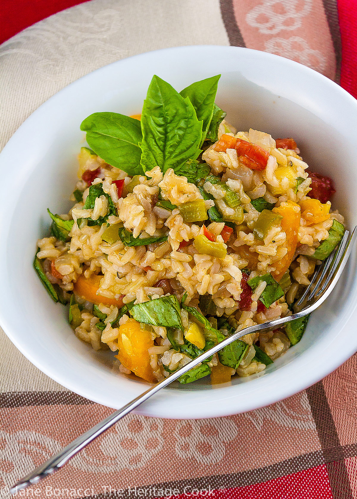 White bowl filled with cooked brown rice tossed with varied vegetables, vinaigrette, and basil leaves; Vegetable Brown Rice Salad © 2023 Jane Bonacci, The Heritage Cook. 