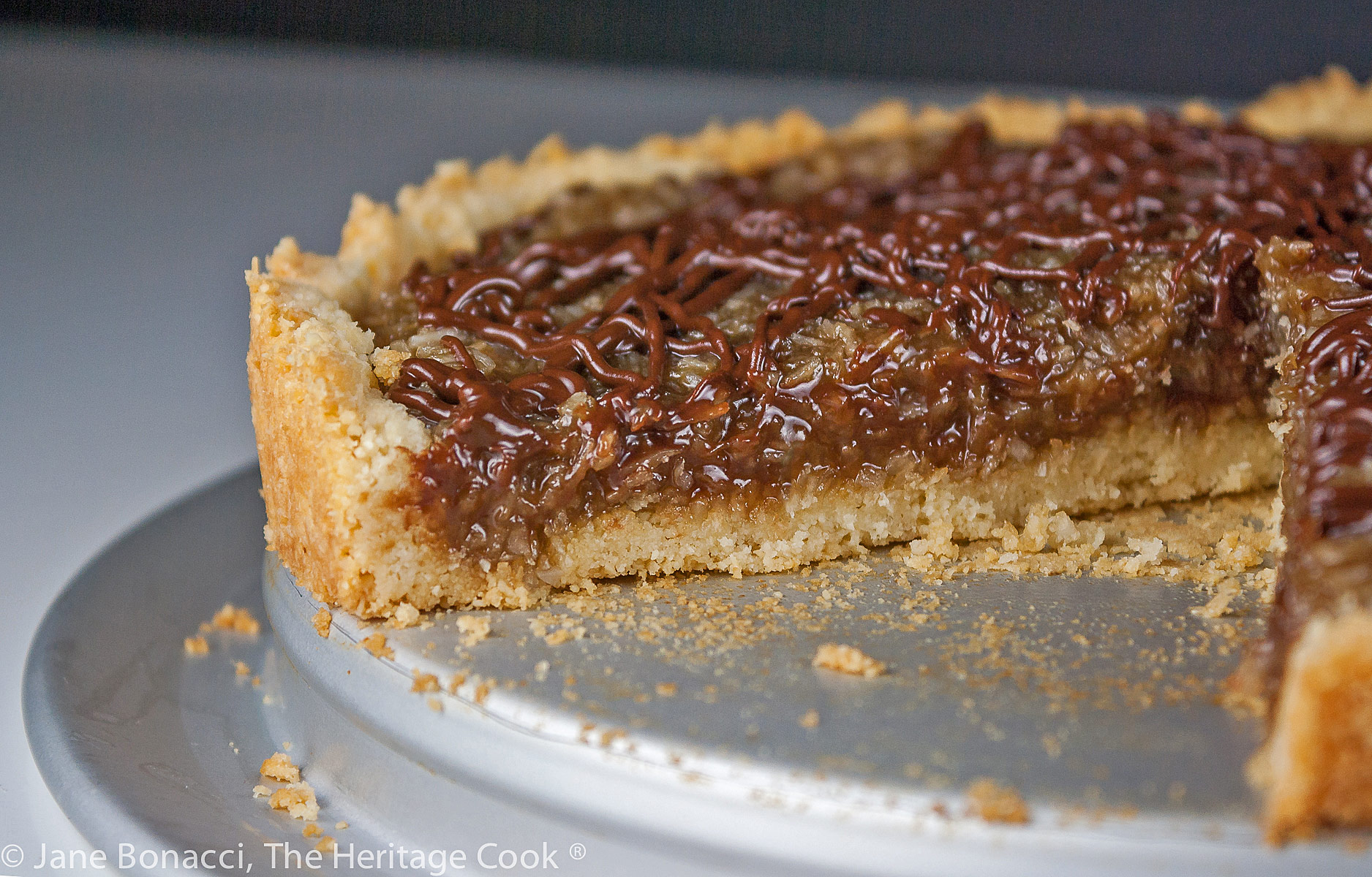 Coconut Caramel Cookie Tart that tastes like a Samoa Girl Scout Cookie; beautiful wedge of the tart drizzled with chocolate and ready to dive into © 2023 Jane Bonacci, The Heritage Cook.
