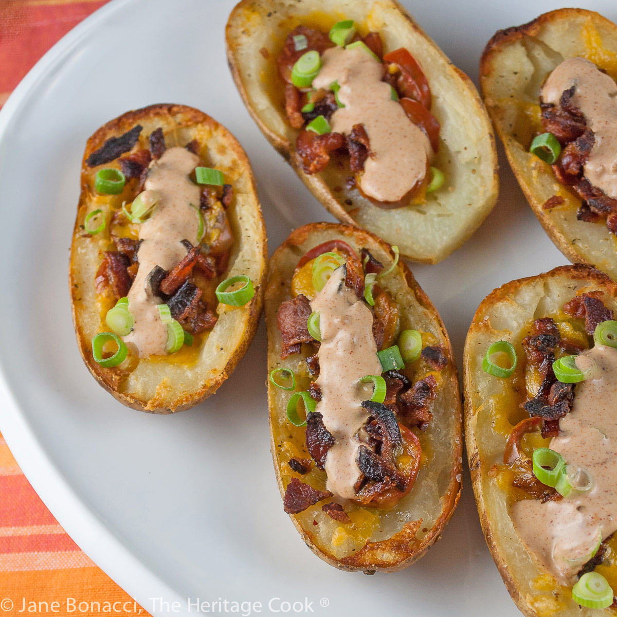 Baked potatoes are split lengthwise with the centers hollowed out and filled with crumbled bacon, green chiles, tomatoes, and melted cheese. Topped with green onions and a drizzle of taco cream. Served with lime wedges. © 2023 Jane Bonacci, The Heritage Cook.