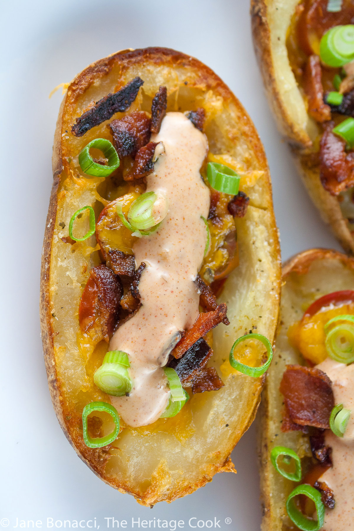 Baked potatoes are split lengthwise with the centers hollowed out and filled with crumbled bacon, green chiles, tomatoes, and melted cheese. Topped with green onions and a drizzle of taco cream. Served with lime wedges. © 2023 Jane Bonacci, The Heritage Cook. 