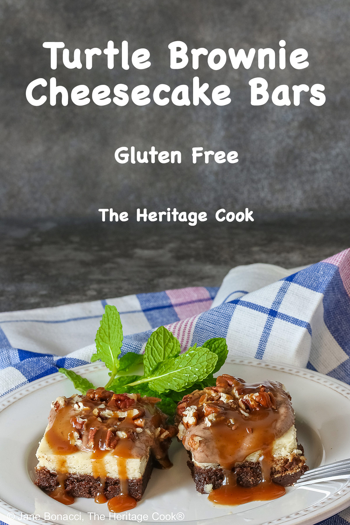 Two Turtle Brownie Cheesecake Bars on a cream colored plate with mint behind them with caramel sauce drizzled over the tops and sitting on a blue, white, and pink checked cloth © 2026 Jane Bonacci, The Heritage Cook.