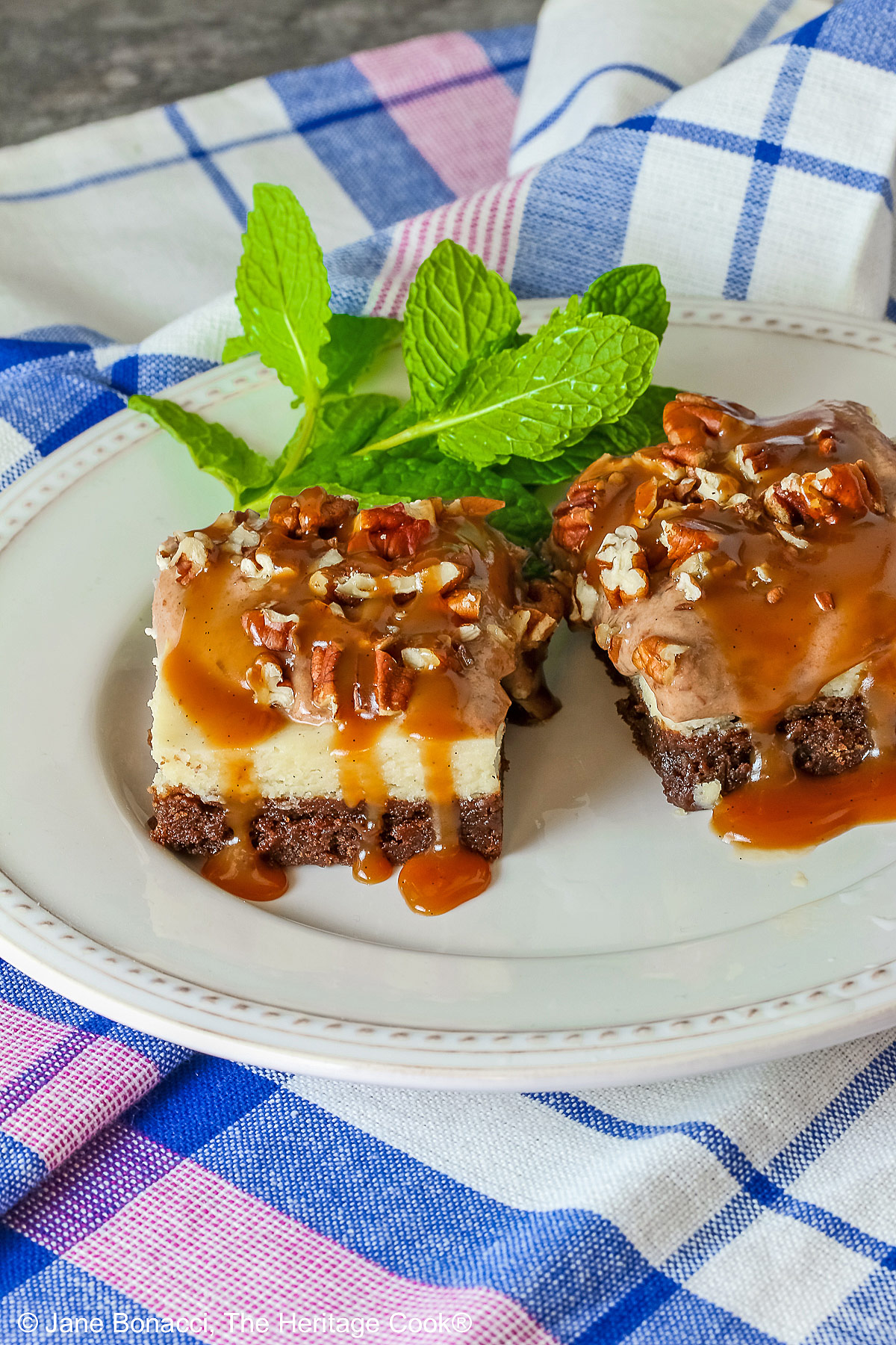 Two Turtle Brownie Cheesecake Bars on a cream colored plate with mint behind them with caramel sauce drizzled over the tops and sitting on a blue, white, and pink checked cloth © 2026 Jane Bonacci, The Heritage Cook.