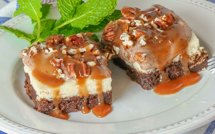 Two Turtle Brownie Cheesecake Bars on a cream colored plate with mint behind them with caramel sauce drizzled over the tops and sitting on a blue, white, and pink checked cloth © 2026 Jane Bonacci, The Heritage Cook.