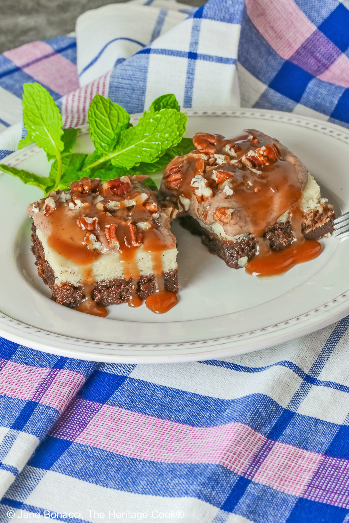 Two Turtle Brownie Cheesecake Bars on a cream colored plate with mint behind them with caramel sauce drizzled over the tops and sitting on a blue, white, and pink checked cloth © 2026 Jane Bonacci, The Heritage Cook.