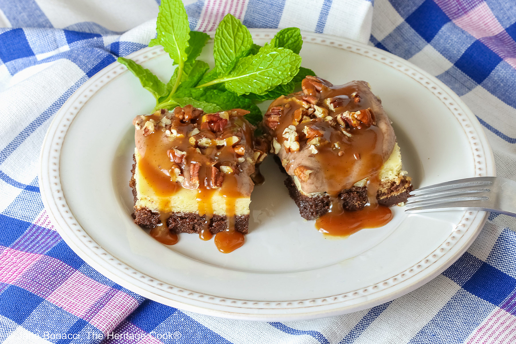 Two Turtle Brownie Cheesecake Bars on a cream colored plate with mint behind them with caramel sauce drizzled over the tops and sitting on a blue, white, and pink checked cloth © 2026 Jane Bonacci, The Heritage Cook.