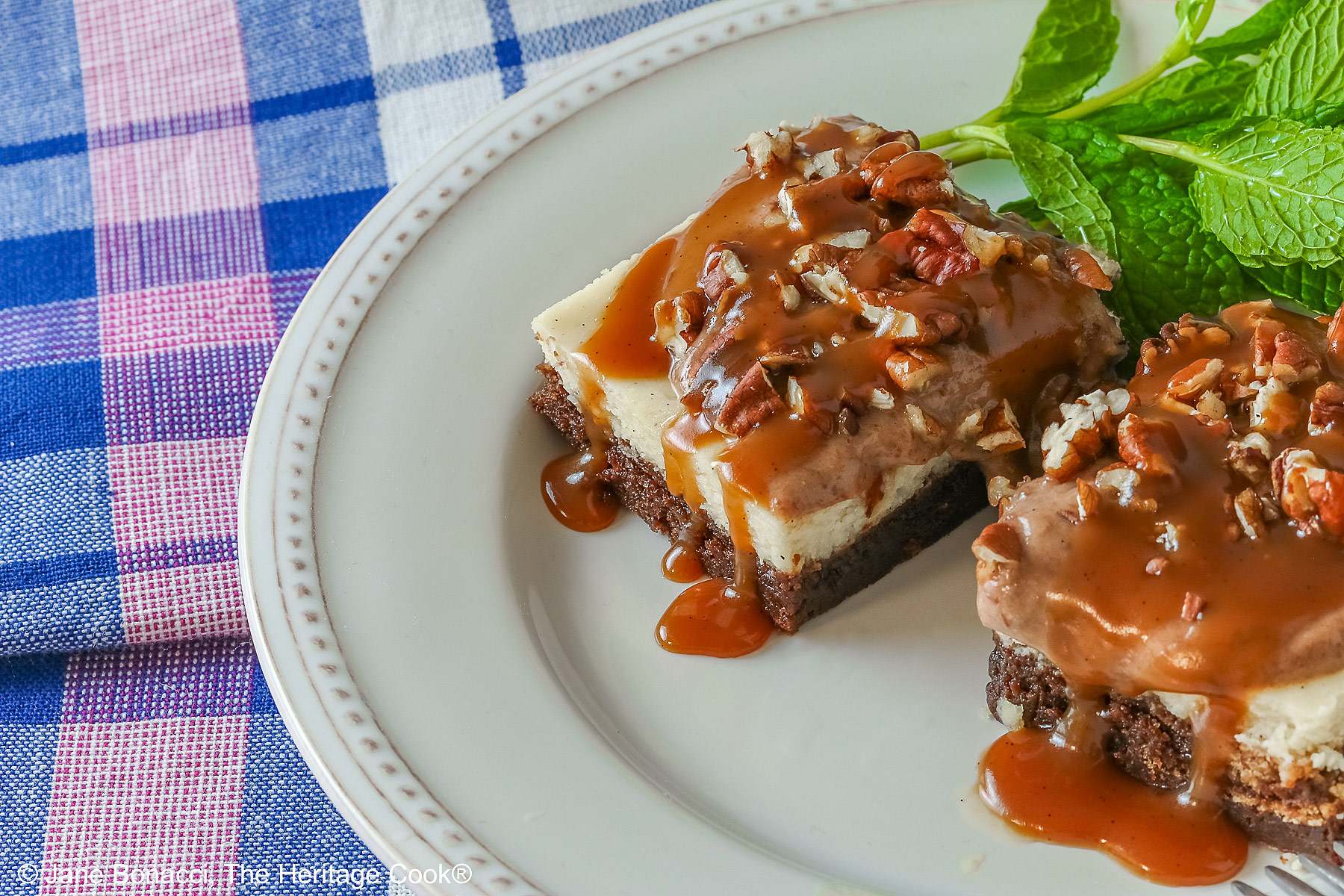 Two Turtle Brownie Cheesecake Bars on a cream colored plate with mint behind them with caramel sauce drizzled over the tops and sitting on a blue, white, and pink checked cloth © 2026 Jane Bonacci, The Heritage Cook.