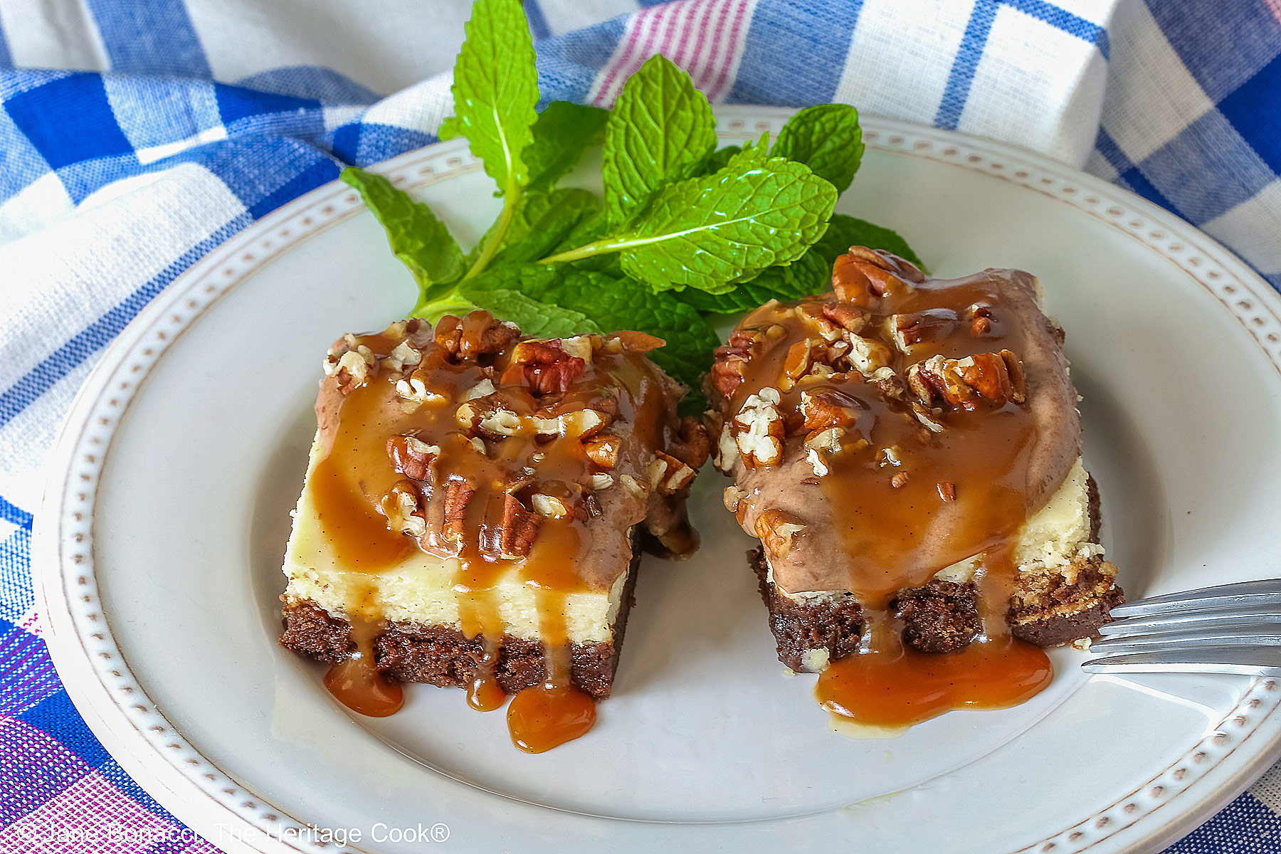 Two Turtle Brownie Cheesecake Bars on a cream colored plate with mint behind them with caramel sauce drizzled over the tops and sitting on a blue, white, and pink checked cloth © 2026 Jane Bonacci, The Heritage Cook.