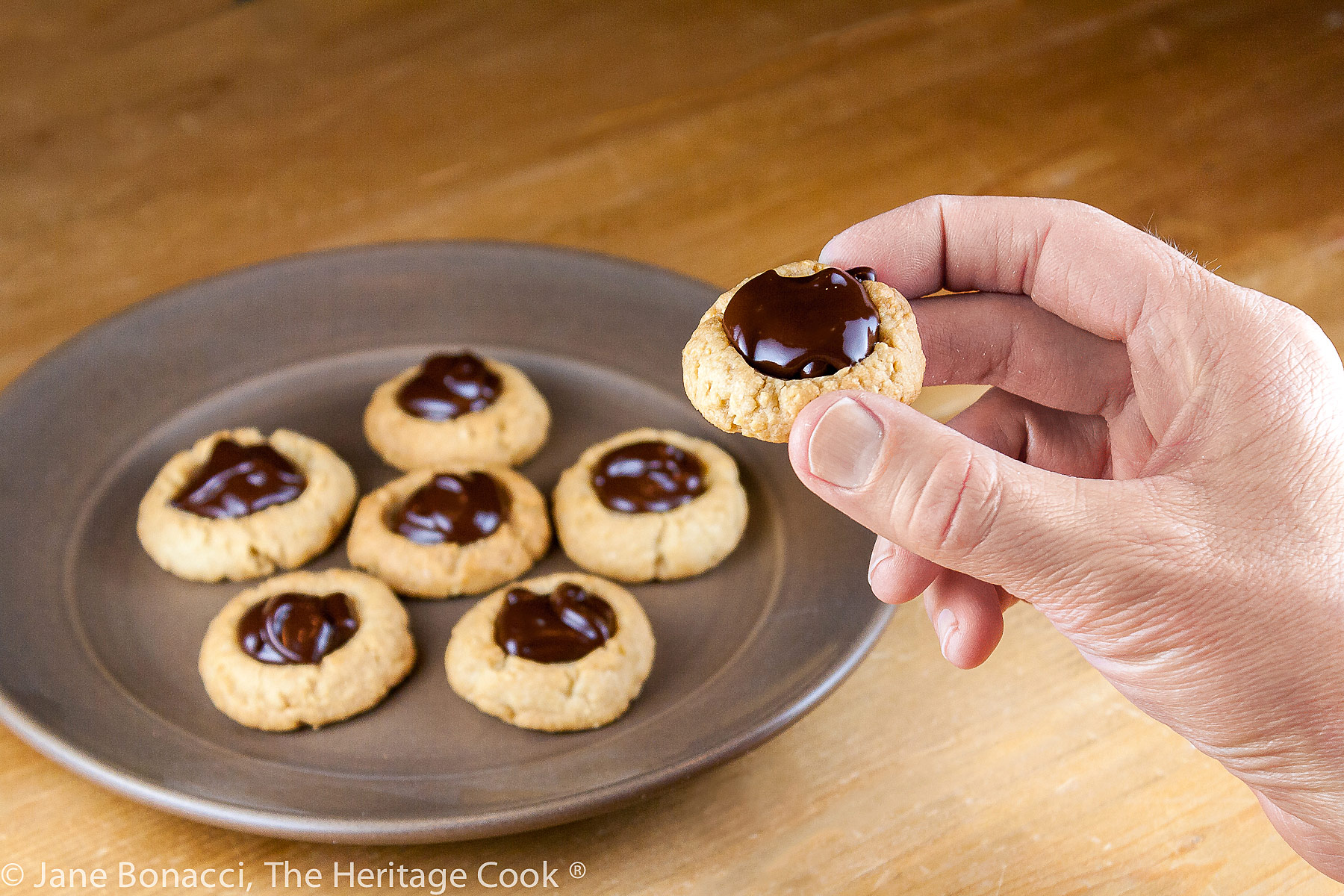 Golden cookies filled with luscious chocolate ganache on a dark brown plate; Almond Chocolate Thumbprint Cookies © 2023 Jane Bonacci, The Heritage Cook. 
