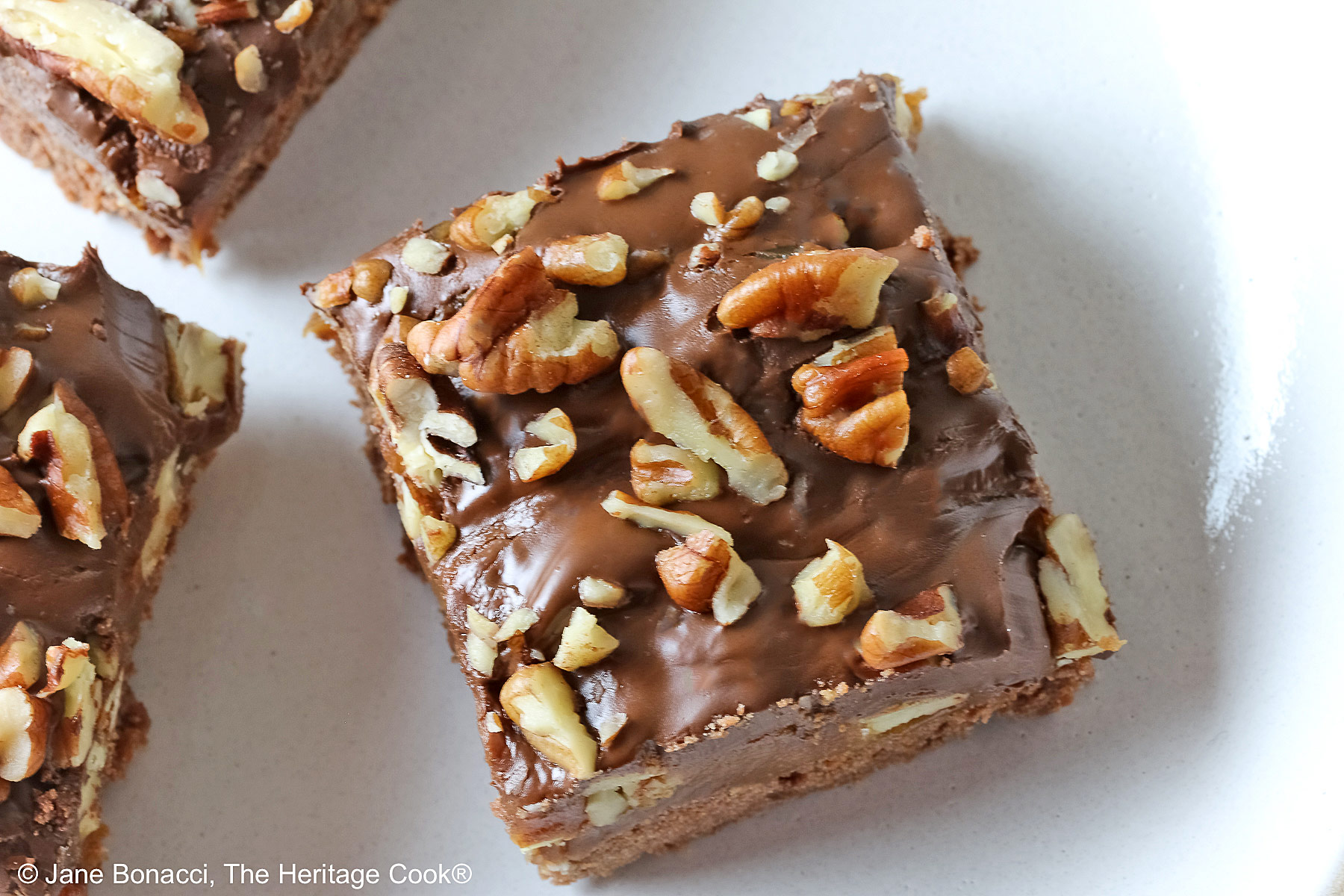 Focus on the top of a single piece. Three Caramel, Pecan, and Chocolate Turtle Bars on a plain white plate sitting on a gray background with a red and white striped cloth beneath the plate or 3 bars stacked offset on a white plate with a blue checked cloth behind © 2026 Jane Bonacci, The Heritage Cook.
