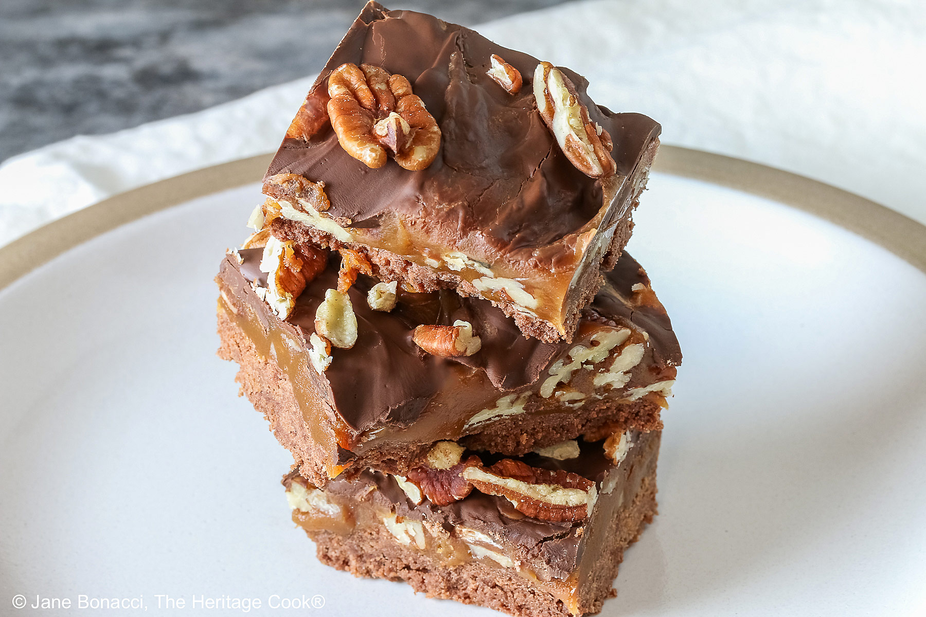 Three Caramel, Pecan, and Chocolate Turtle Bars on a plain white plate sitting on a gray background with a red and white striped cloth beneath the plate or 3 bars stacked offset on a white plate with a blue checked cloth behind © 2026 Jane Bonacci, The Heritage Cook.