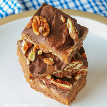 Three Caramel, Pecan, and Chocolate Turtle Bars on a plain white plate sitting on a gray background with a red and white striped cloth beneath the plate or 3 bars stacked offset on a white plate with a blue checked cloth behind © 2026 Jane Bonacci, The Heritage Cook.