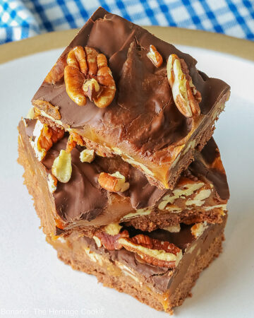 Three Caramel, Pecan, and Chocolate Turtle Bars on a plain white plate sitting on a gray background with a red and white striped cloth beneath the plate or 3 bars stacked offset on a white plate with a blue checked cloth behind © 2026 Jane Bonacci, The Heritage Cook.