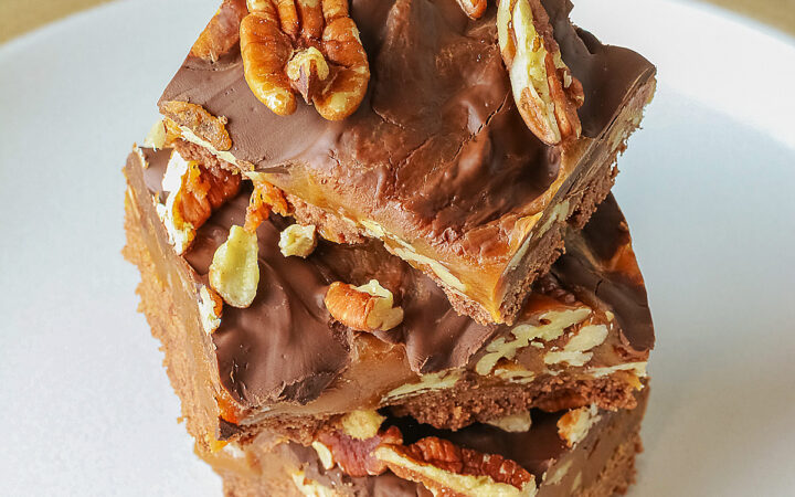 Three Caramel, Pecan, and Chocolate Turtle Bars on a plain white plate sitting on a gray background with a red and white striped cloth beneath the plate or 3 bars stacked offset on a white plate with a blue checked cloth behind © 2026 Jane Bonacci, The Heritage Cook.
