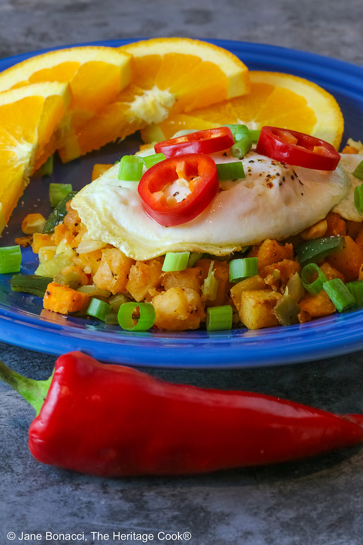 A large scoop of the Southwest Breakfast Hash on a dark blue plate topped with a fried egg, red Fresno chile slices, and chopped green onions, flanked by beautiful orange slices and served with salsa on the side © 2025 Jane Bonacci, The Heritage Cook.