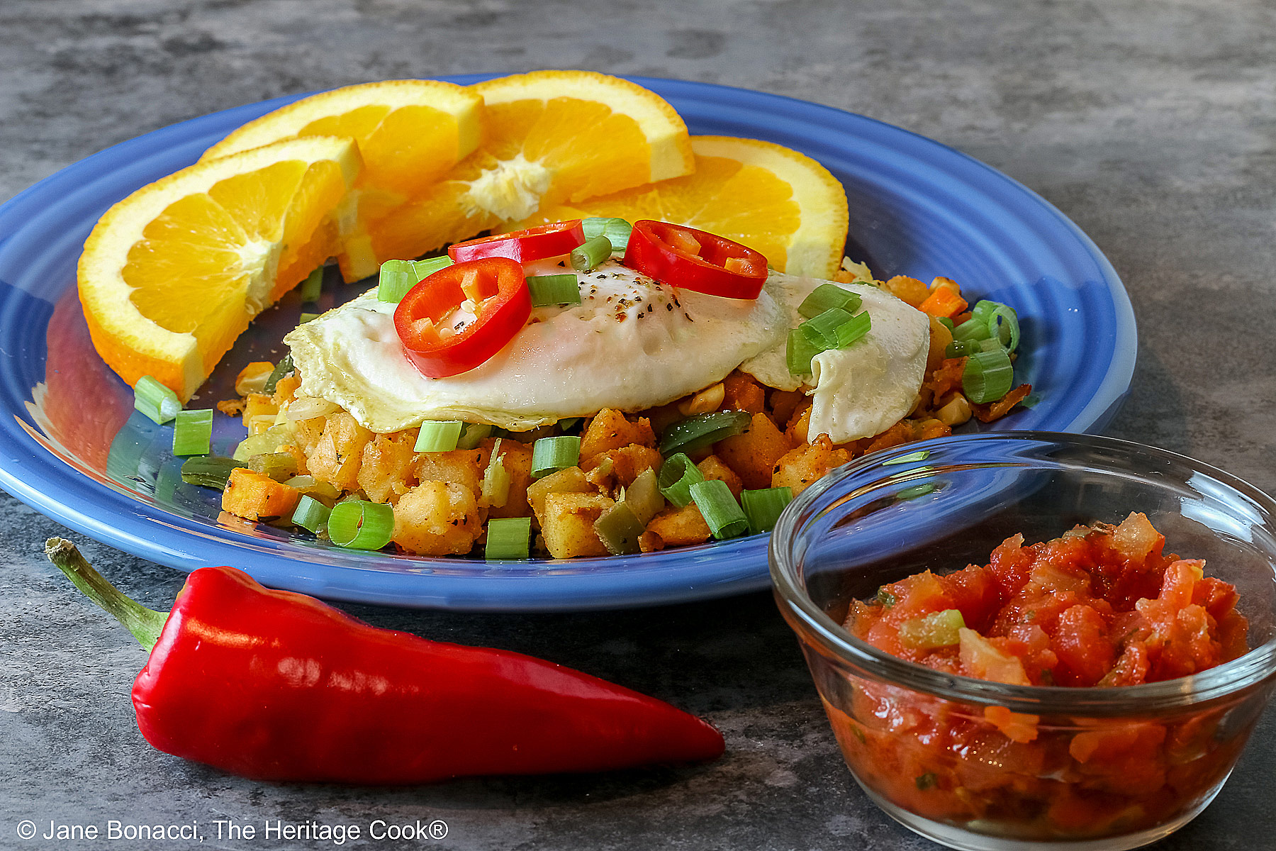 A large scoop of the Southwest Breakfast Hash on a dark blue plate topped with a fried egg, red Fresno chile slices, and chopped green onions, flanked by beautiful orange slices and served with salsa on the side © 2025 Jane Bonacci, The Heritage Cook.