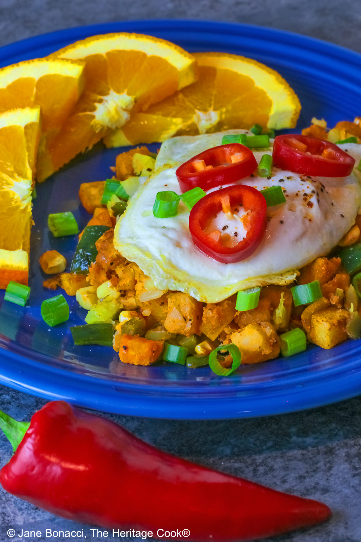 A large scoop of the Southwest Breakfast Hash on a dark blue plate topped with a fried egg, red Fresno chile slices, and chopped green onions, flanked by beautiful orange slices and served with salsa on the side © 2025 Jane Bonacci, The Heritage Cook.