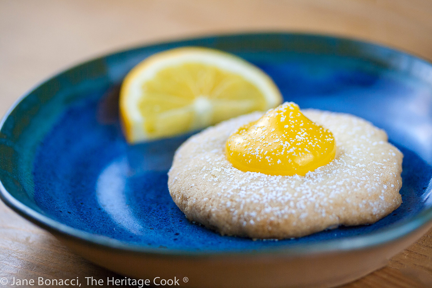 Pale golden shortbread cookies filled with a dollop of bright yellow Meyer lemon curd, on blue plates or wire racks, some sprinkled with confectioners’ sugar © 2024 Jane Bonacci, The Heritage Cook. 