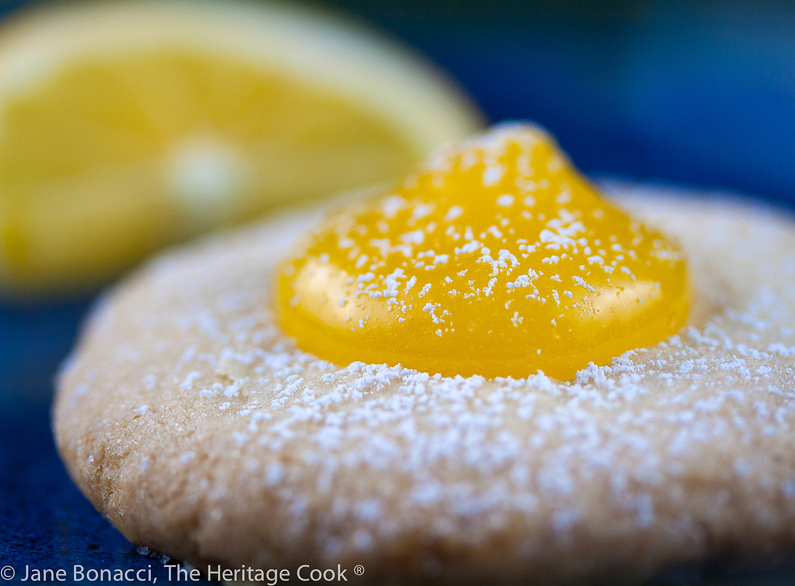 Pale golden shortbread cookies filled with a dollop of bright yellow Meyer lemon curd, on blue plates or wire racks, some sprinkled with confectioners’ sugar © 2024 Jane Bonacci, The Heritage Cook. 
