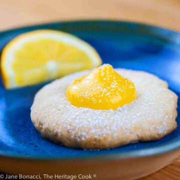 Pale golden shortbread cookies filled with a dollop of bright yellow Meyer lemon curd, on blue plates or wire racks, some sprinkled with confectioners’ sugar © 2024 Jane Bonacci, The Heritage Cook.
