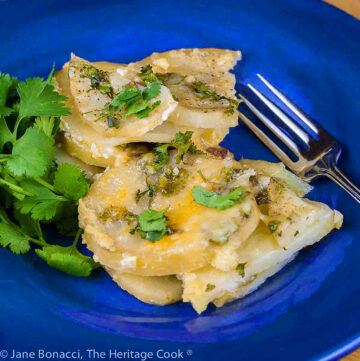Slices of potatoes covered with melted cheese and dotted with green chiles, sprinkled with chopped cilantro on deep blue bowl © 2024 Jane Bonacci, The Heritage Cook.