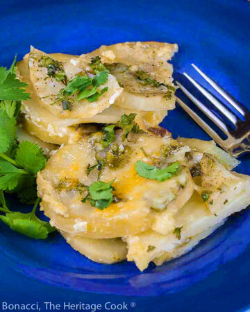 Slices of potatoes covered with melted cheese and dotted with green chiles, sprinkled with chopped cilantro on deep blue bowl © 2024 Jane Bonacci, The Heritage Cook.