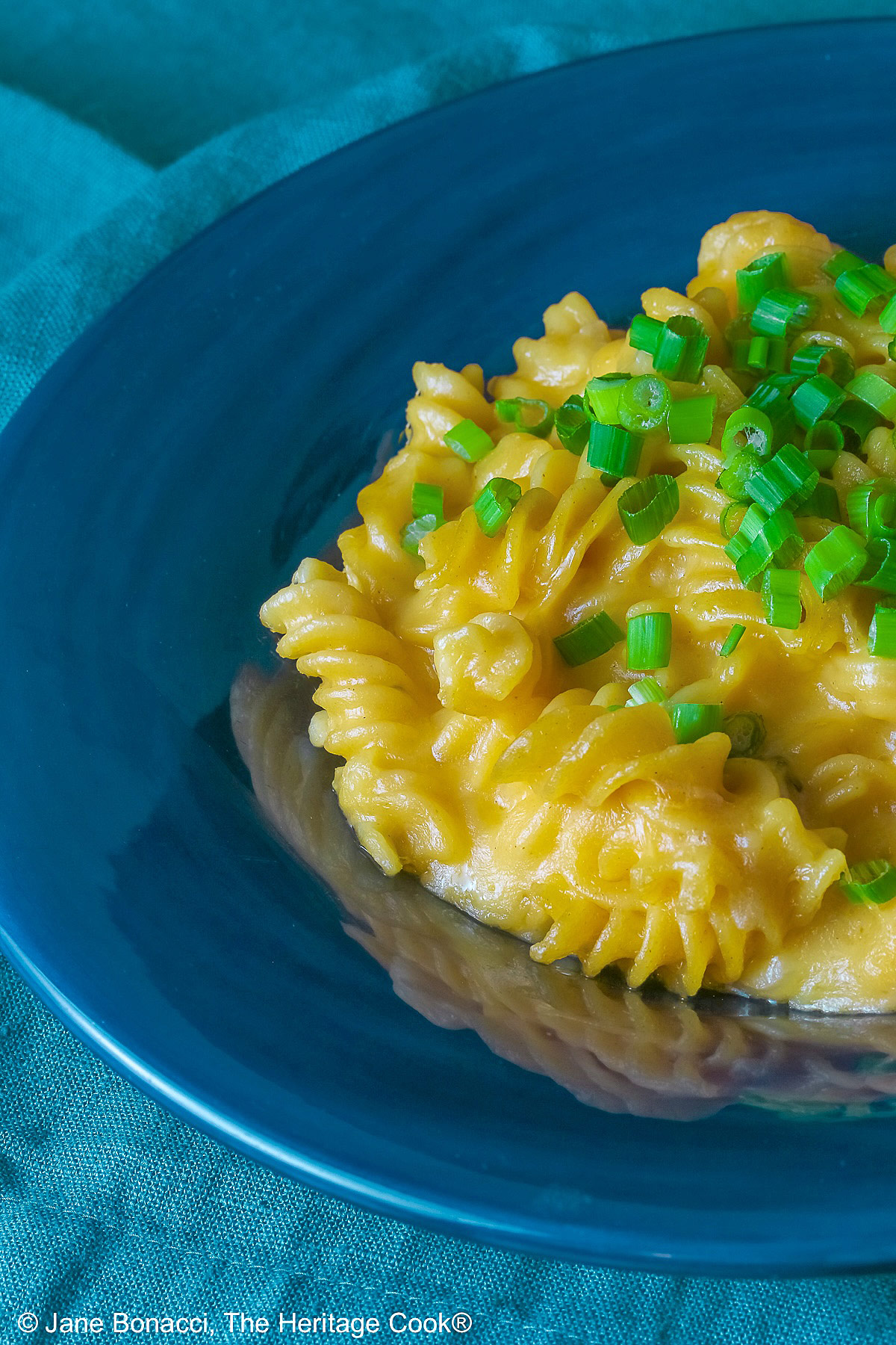 Wide dark blue bowl with a big rim filled with golden Swiss Fondue Mac and 4 Cheese, topped with a lot of sliced green onions, some with a fork in the back, sitting on an aqua cloth © 2026 Jane Bonacci, The Heritage Cook.