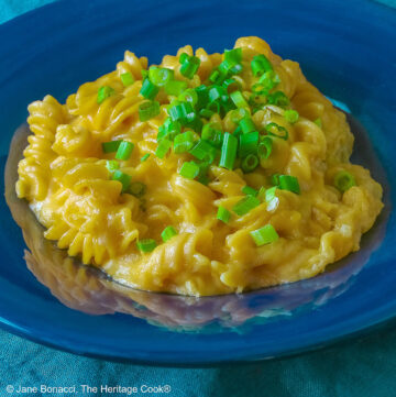 Wide dark blue bowl with a big rim filled with golden Swiss Fondue Mac and 4 Cheese, topped with a lot of sliced green onions, some with a fork in the back, sitting on an aqua cloth © 2026 Jane Bonacci, The Heritage Cook.