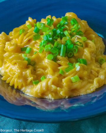 Wide dark blue bowl with a big rim filled with golden Swiss Fondue Mac and 4 Cheese, topped with a lot of sliced green onions, some with a fork in the back, sitting on an aqua cloth © 2026 Jane Bonacci, The Heritage Cook.