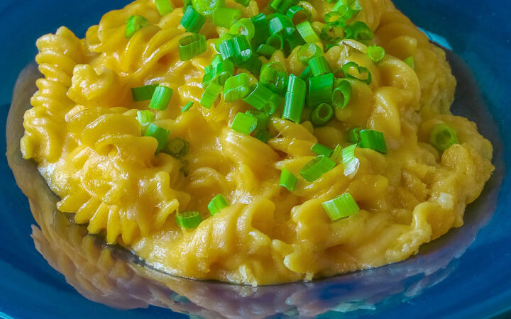 Wide dark blue bowl with a big rim filled with golden Swiss Fondue Mac and 4 Cheese, topped with a lot of sliced green onions, some with a fork in the back, sitting on an aqua cloth © 2026 Jane Bonacci, The Heritage Cook.