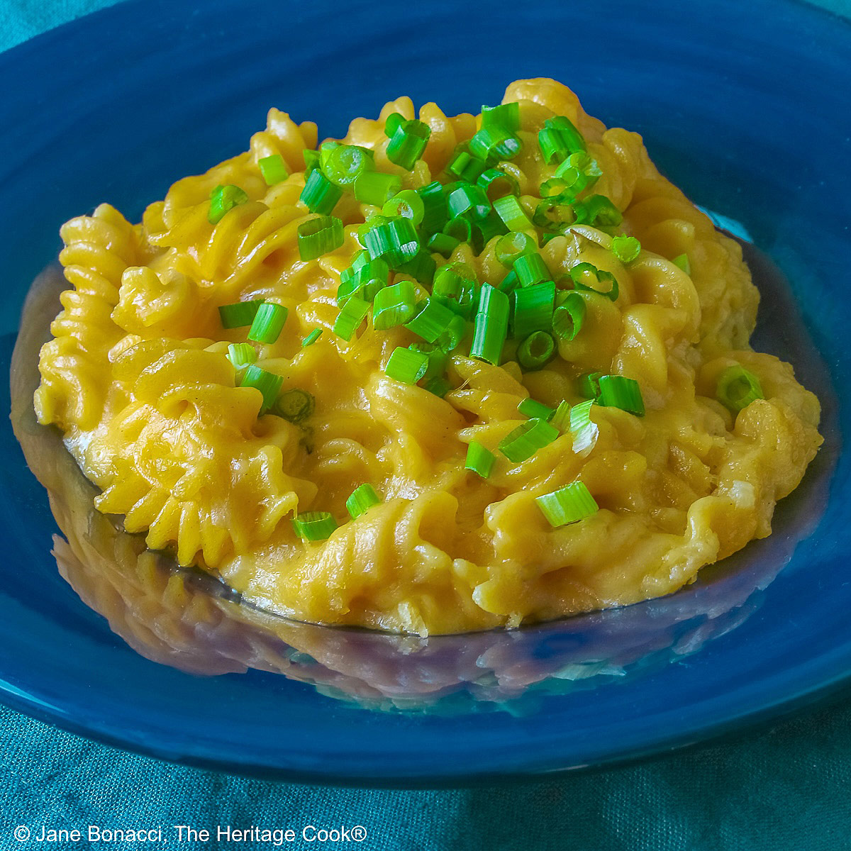 Wide dark blue bowl with a big rim filled with golden Swiss Fondue Mac and 4 Cheese, topped with a lot of sliced green onions, some with a fork in the back, sitting on an aqua cloth © 2026 Jane Bonacci, The Heritage Cook.