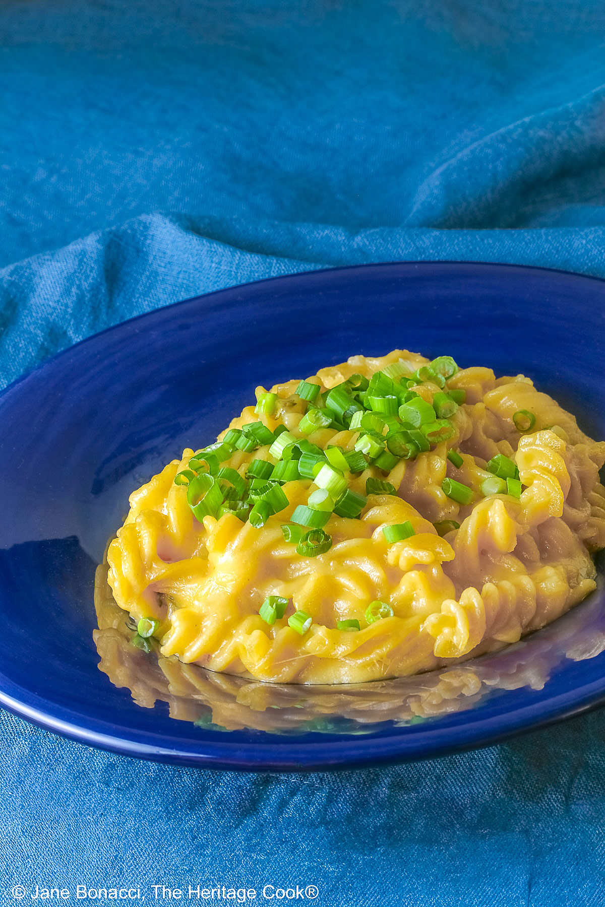 Wide dark blue bowl with a big rim filled with golden Swiss Fondue Mac and 4 Cheese, topped with a lot of sliced green onions, some with a fork in the back, sitting on an aqua cloth © 2026 Jane Bonacci, The Heritage Cook.