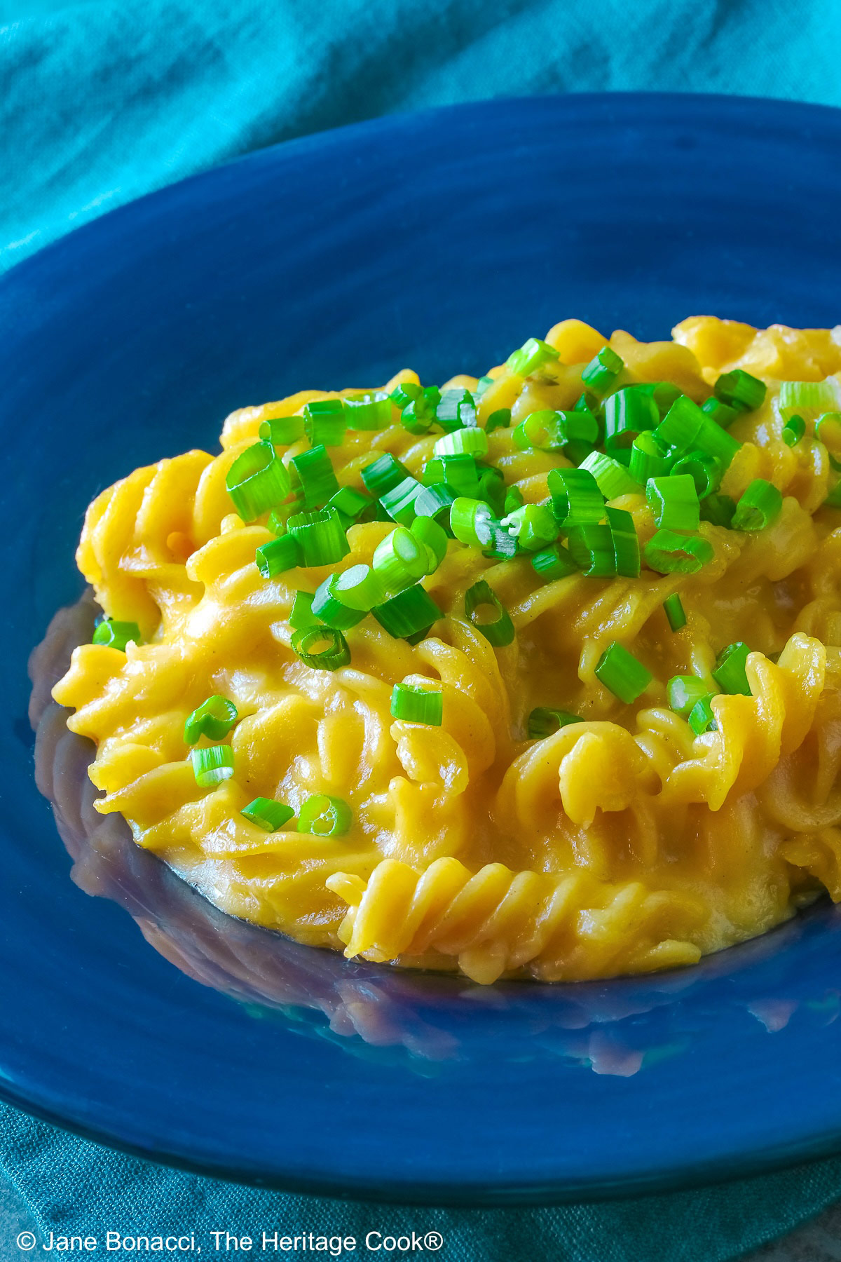 Wide dark blue bowl with a big rim filled with golden Swiss Fondue Mac and 4 Cheese, topped with a lot of sliced green onions, some with a fork in the back, sitting on an aqua cloth © 2026 Jane Bonacci, The Heritage Cook.
