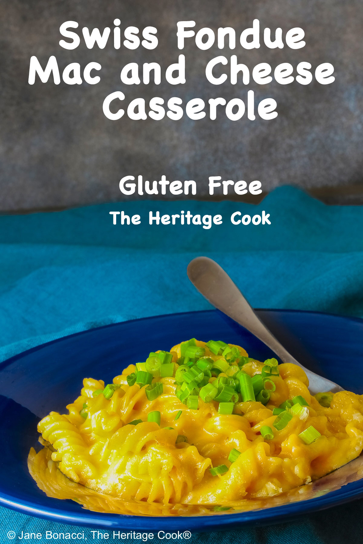 Wide dark blue bowl with a big rim filled with golden Swiss Fondue Mac and 4 Cheese, topped with a lot of sliced green onions, some with a fork in the back, sitting on an aqua cloth © 2026 Jane Bonacci, The Heritage Cook.