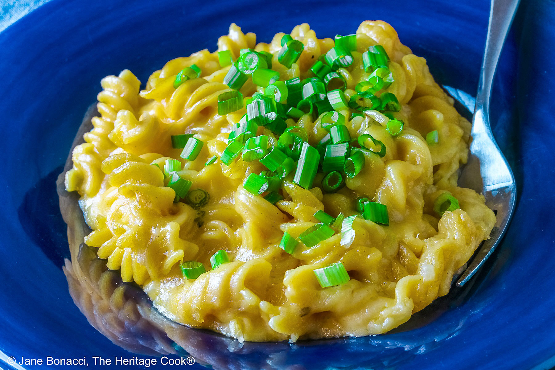 Wide dark blue bowl with a big rim filled with golden Swiss Fondue Mac and 4 Cheese, topped with a lot of sliced green onions, some with a fork in the back, sitting on an aqua cloth © 2026 Jane Bonacci, The Heritage Cook.