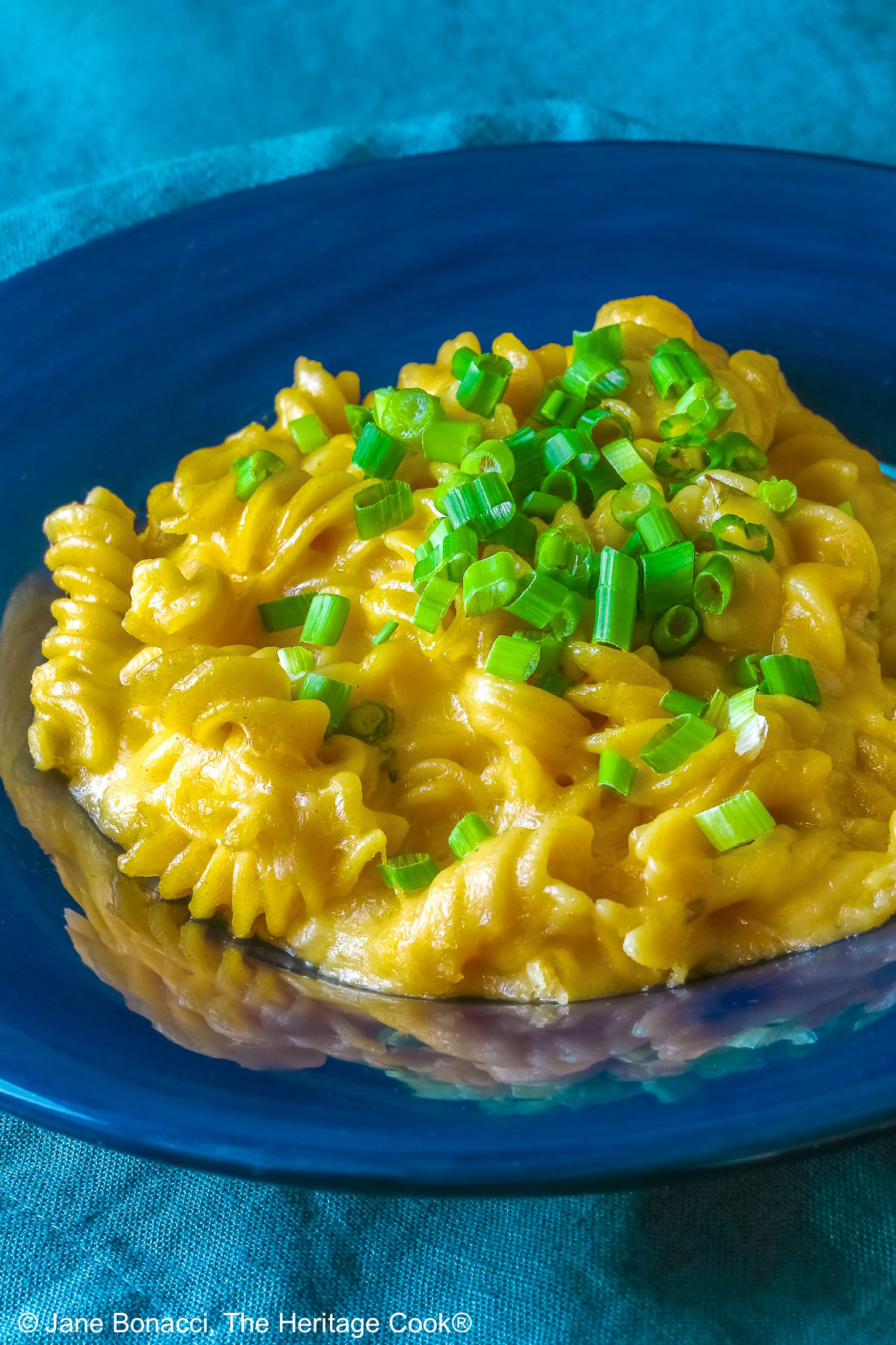 Wide dark blue bowl with a big rim filled with golden Swiss Fondue Mac and 4 Cheese, topped with a lot of sliced green onions, some with a fork in the back, sitting on an aqua cloth © 2026 Jane Bonacci, The Heritage Cook.