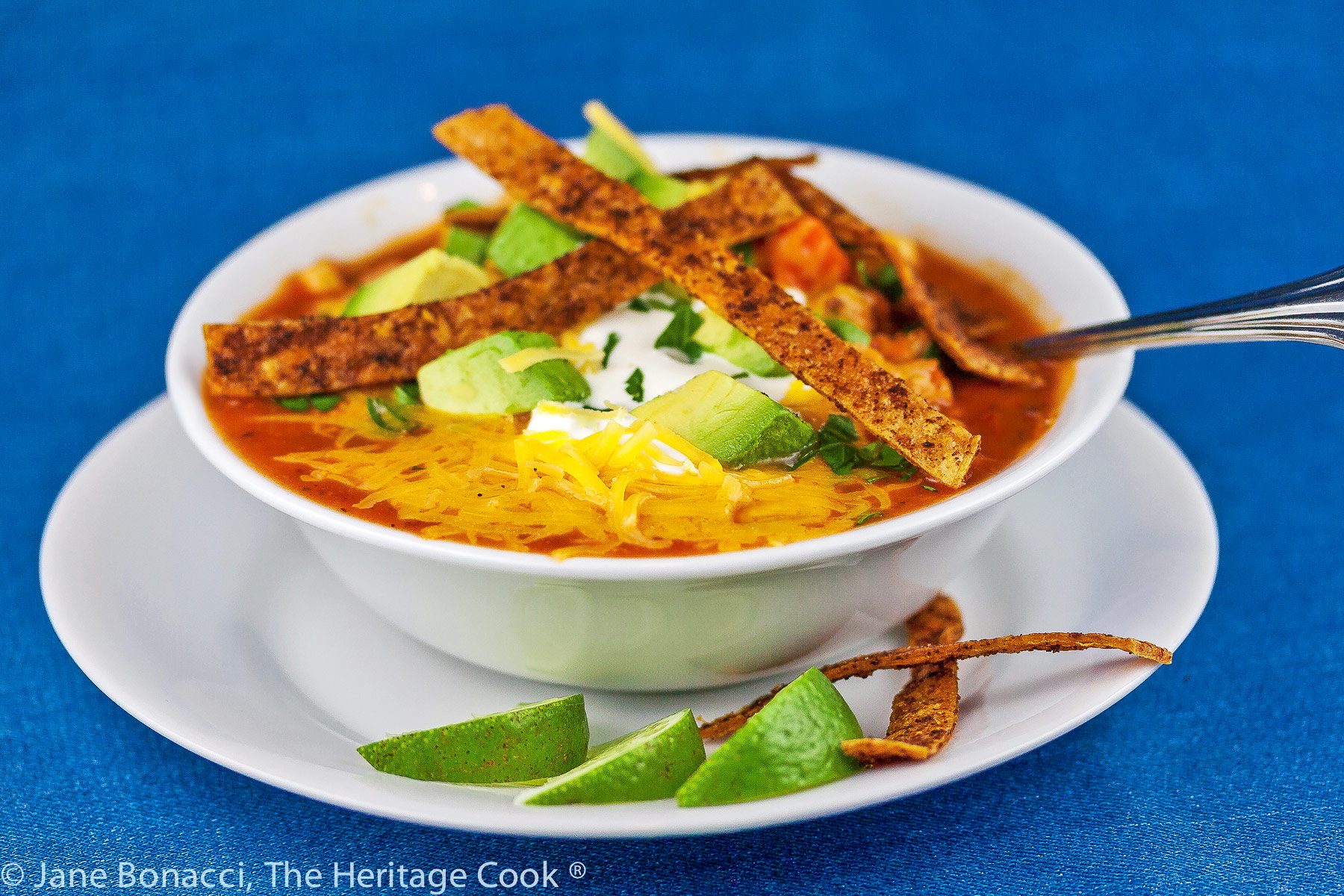 White bowl filled with chile red broth with melted cheese, sour cream, avocados, and spiced tortilla strips on a white plate with lime wedges and more tortilla strips © 2024 Jane Bonacci, The Heritage Cook. 