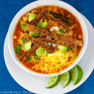 White bowl filled with chile red broth with melted cheese, sour cream, avocados, and spiced tortilla strips on a white plate with lime wedges and more tortilla strips © 2024 Jane Bonacci, The Heritage Cook.