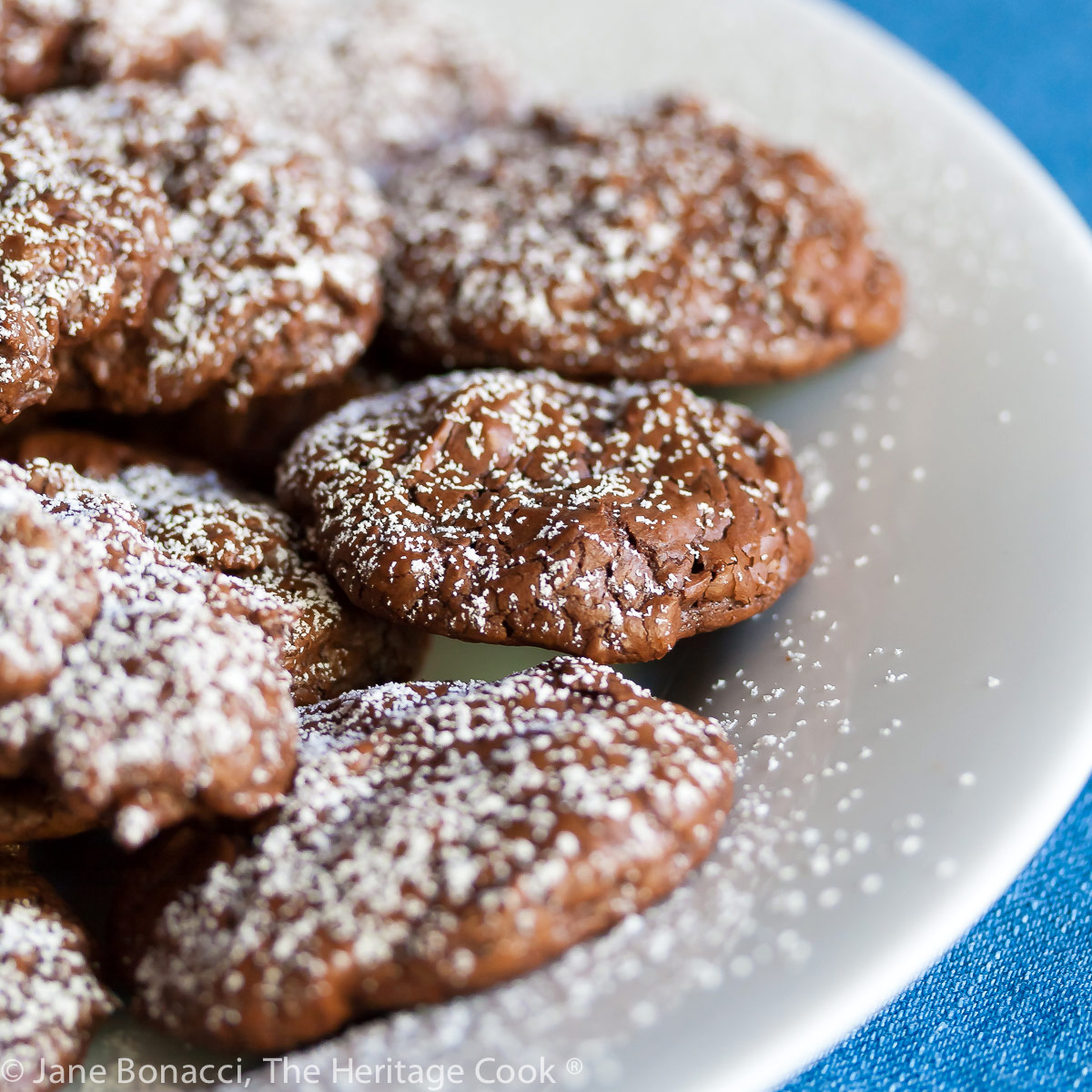 Chocolate Brownie Cookies on a white plate with a bright blue backdrop, some sprinkled with powdered sugar; © 2023 Jane Bonacci, The Heritage Cook. 